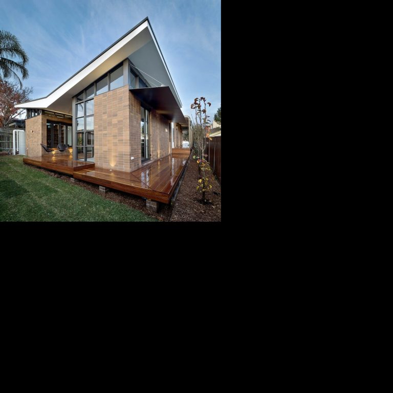 Modern house with large windows, wooden deck, and a sloped roof in a garden setting.