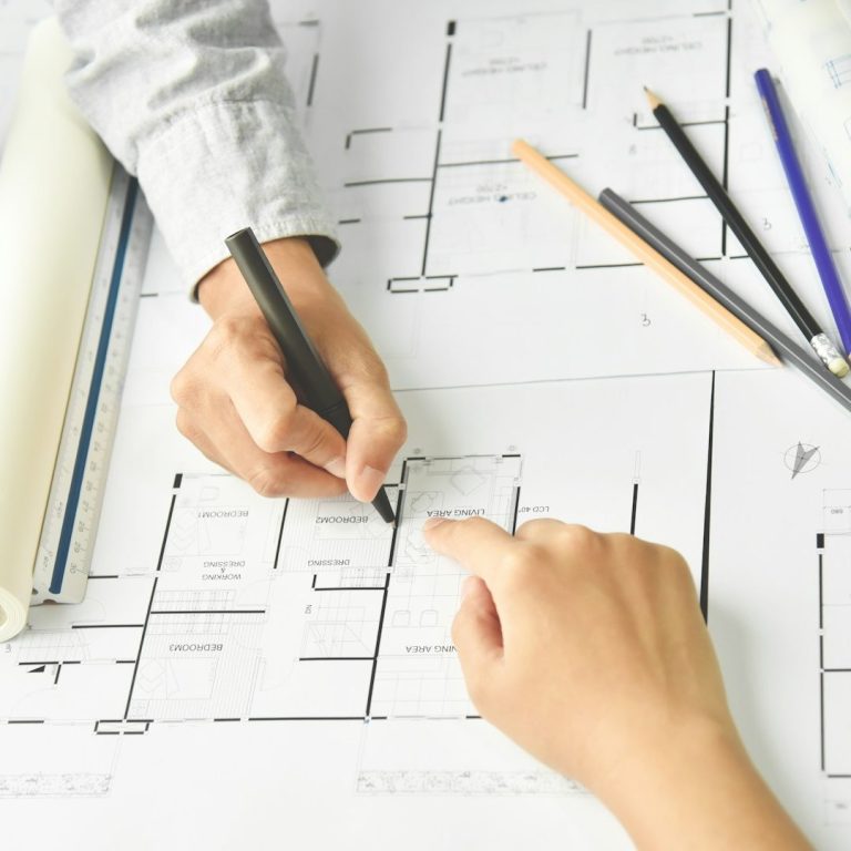 Hands sketching on architectural blueprints with pens and a ruler.
