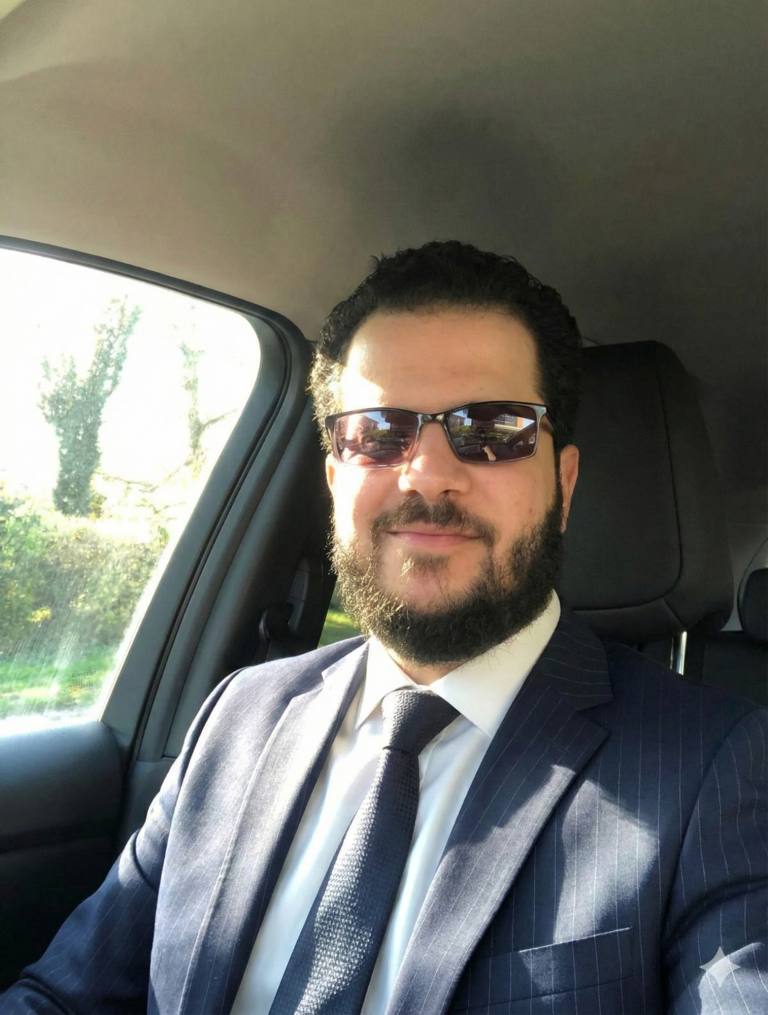 Haitham Elkashawy Man in a suit and sunglasses seated in a car, smiling.