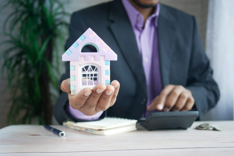 A person in a suit holds a model house while using a calculator on a desk.