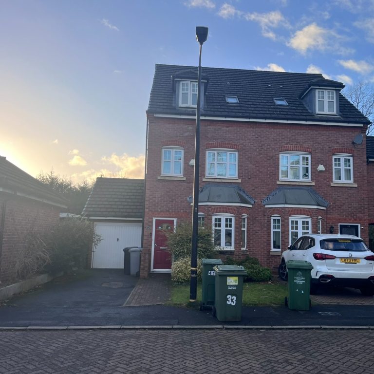 Red-brick house with multiple windows and a white car parked outside, near green bins.