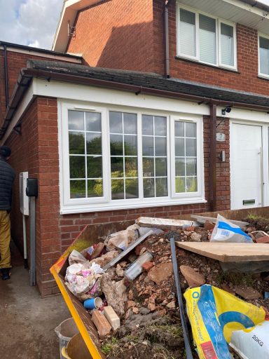 A construction site with a pile of debris in front of a brick house and large windows.