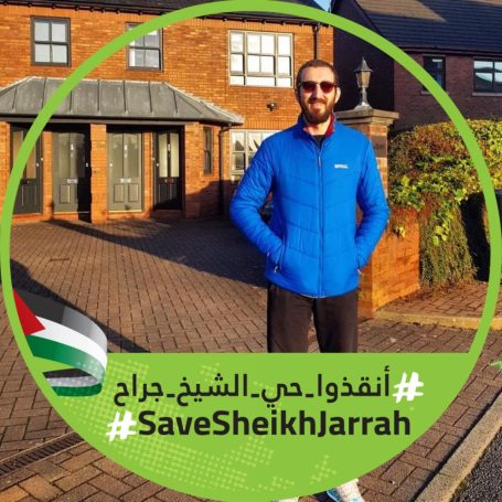 Man in a blue jacket standing outside a brick building, holding a sign about Sheikh Jarrah.